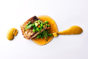 Plato gourmet con pescado, guisantes y salsa en un fondo blanco.