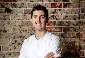 Josh Niland, chef australiano, sonriente frente a una pared de ladrillos.