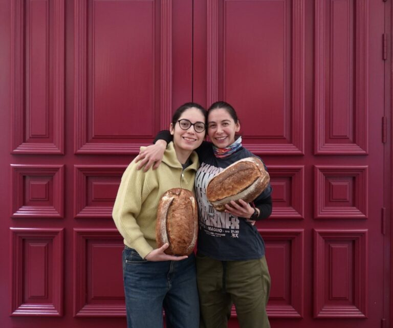 Dos mujeres sosteniendo hogazas de pan frente a una puerta roja