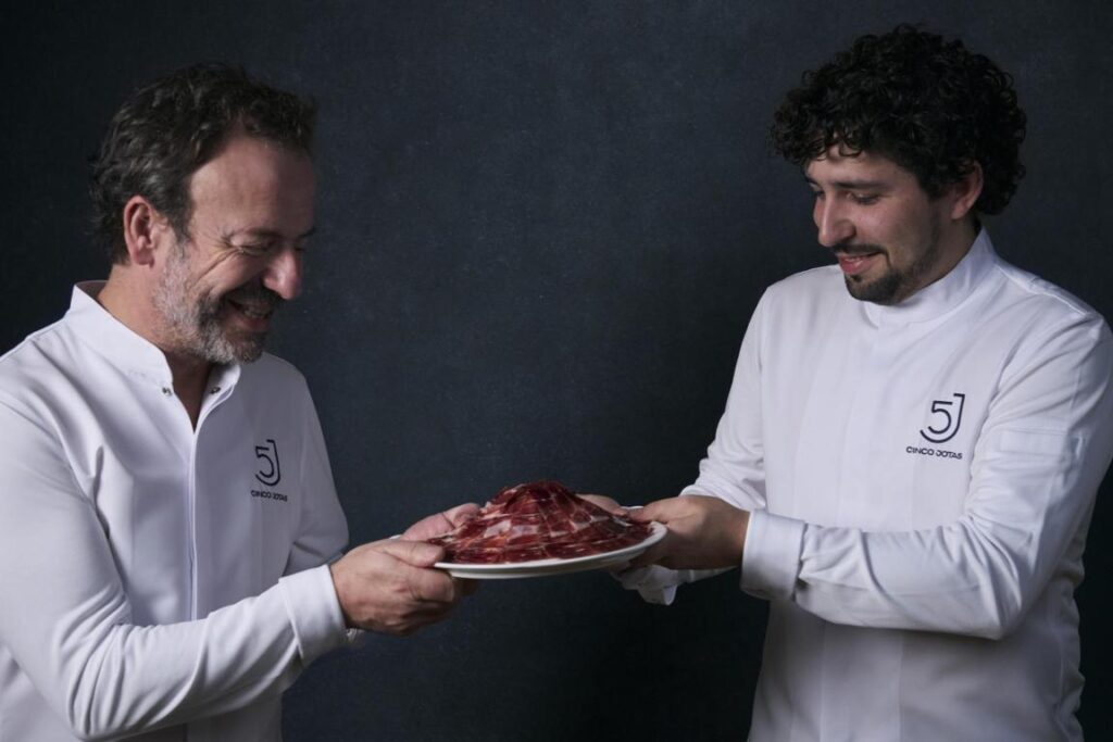 Dos chefs sosteniendo un plato con jamón ibérico en un fondo oscuro.