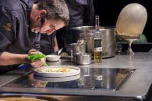 Chef preparando un plato con ingredientes frescos y vino en una cocina profesional.