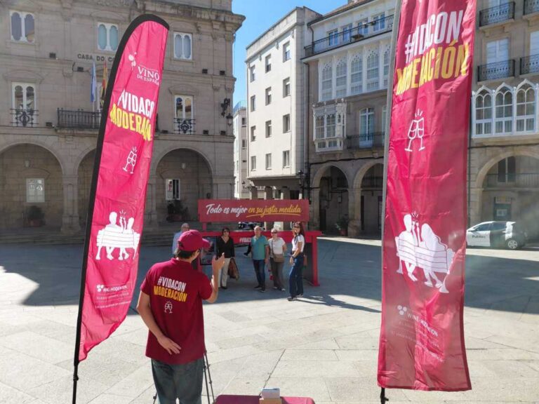 Personas junto a un banco rojo gigante y banderolas de una campaña en una plaza.