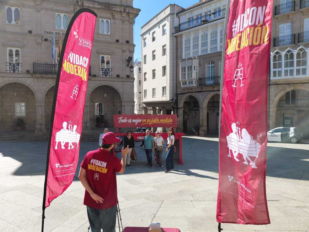 Personas junto a un banco rojo gigante y banderolas de una campaña en una plaza.