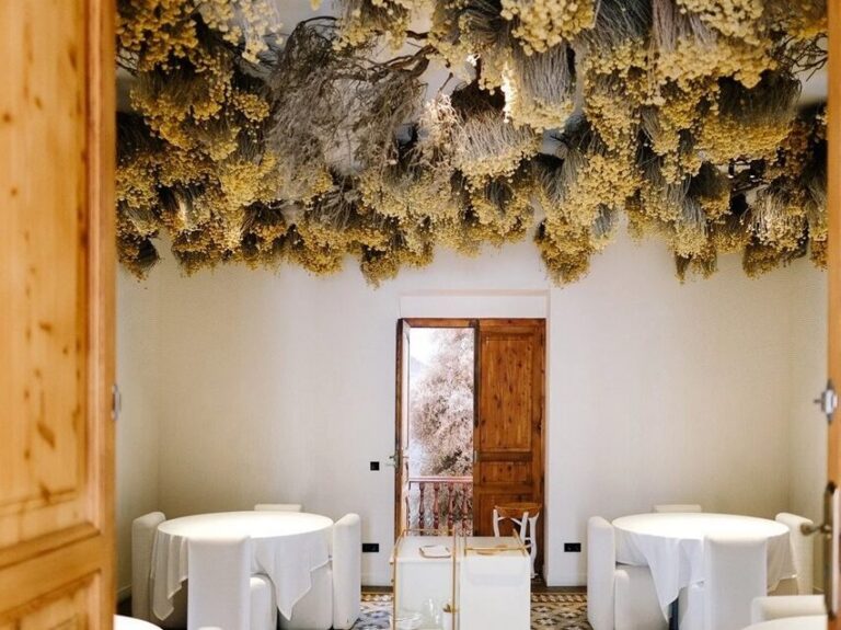 Comedor con mesas redondas vestidas de blanco y plantas secas suspendidas del techo.