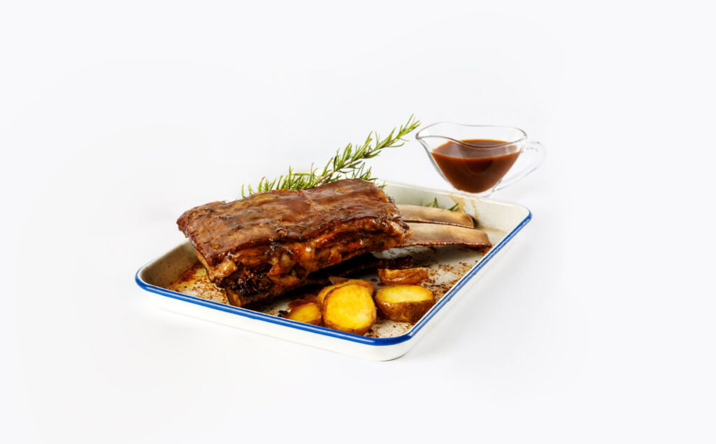 Costilla asada con patatas en una fuente, acompañada de salsa y romero.