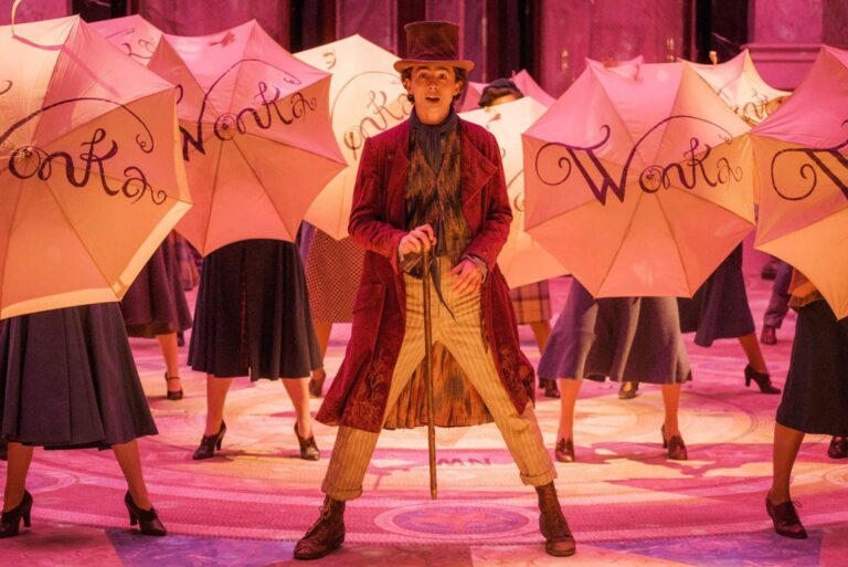 Un hombre con sombrero y abrigo rojo posa entre bailarines con paraguas de Wonka.