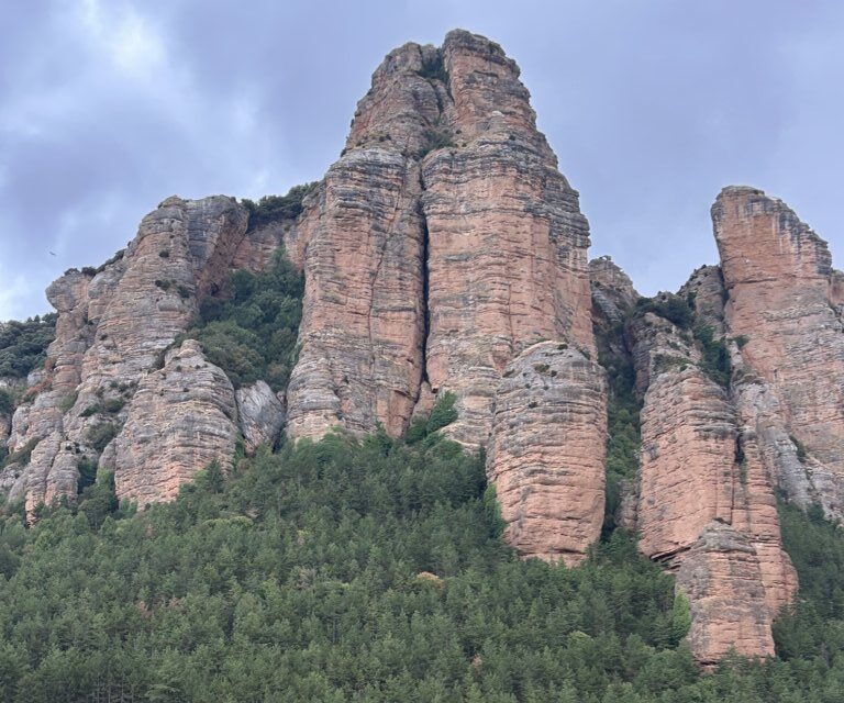 Altas formaciones rocosas rojizas sobre un bosque, con viviendas y parcelas en primer plano.