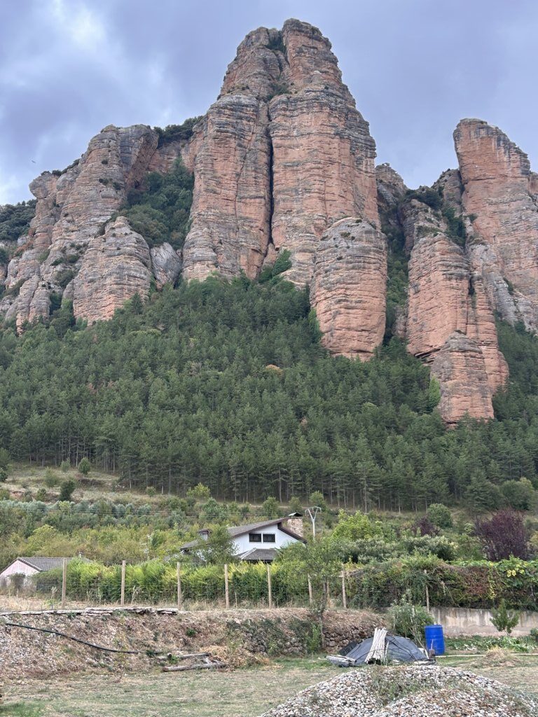 Altas formaciones rocosas rojizas sobre un bosque, con viviendas y parcelas en primer plano.