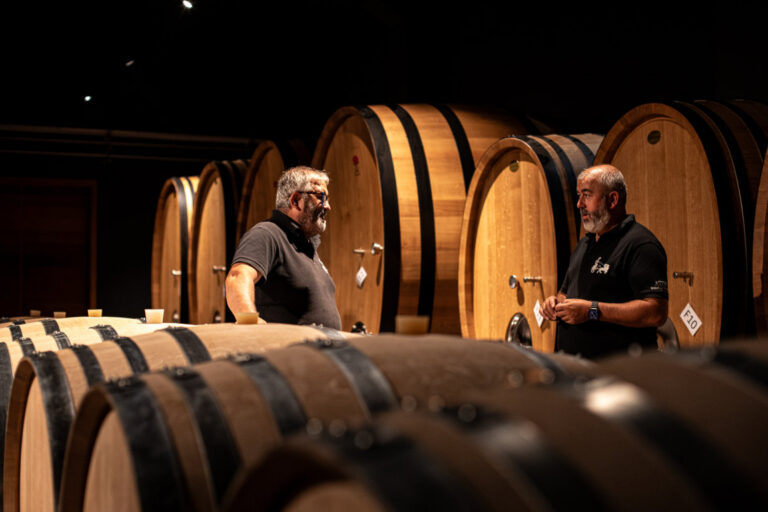 Dos hombres conversando en una bodega con barricas de vino