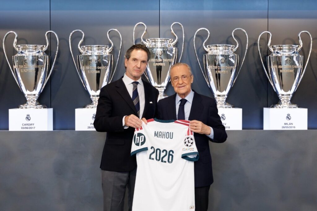 Dos hombres sostienen una camiseta blanca de Mahou frente a varias copas plateadas.