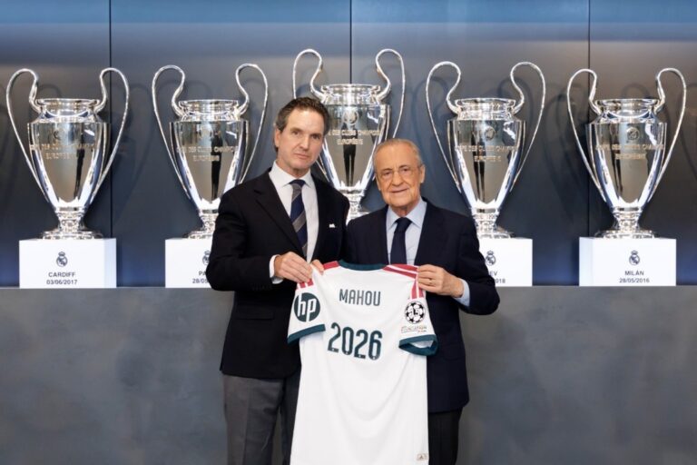 Dos hombres sostienen una camiseta blanca de Mahou frente a varias copas plateadas.