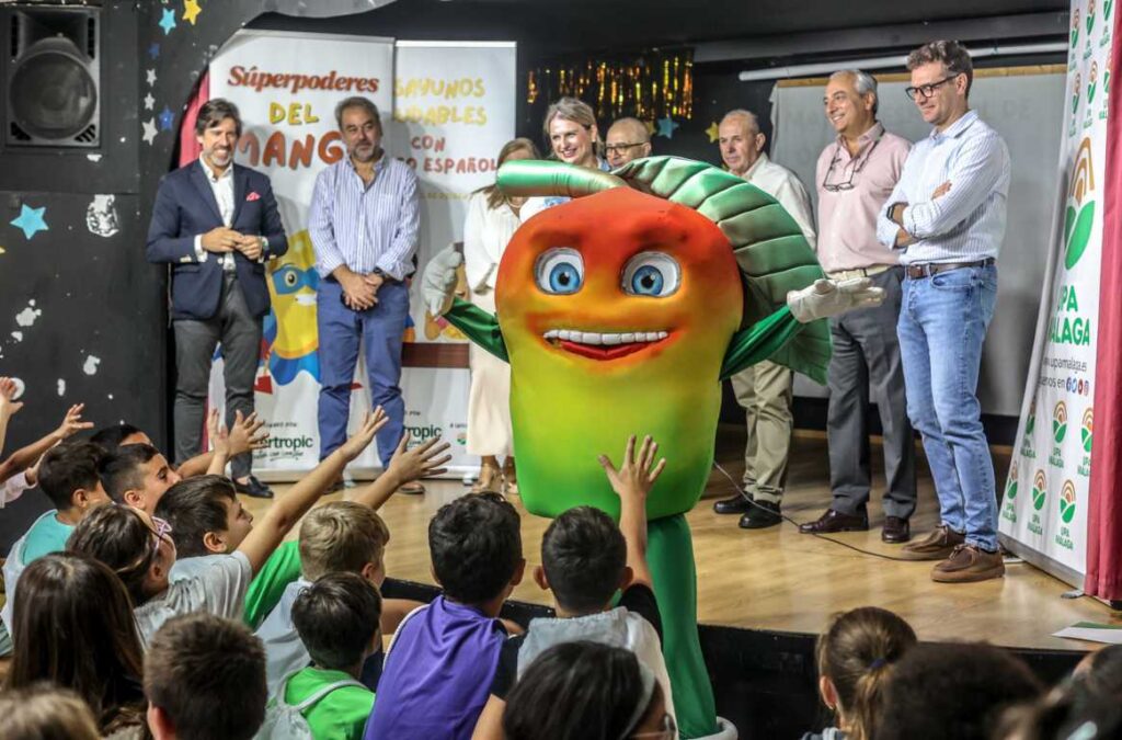Escolares observan a una mascota con forma de mango junto a varios adultos en un escenario.