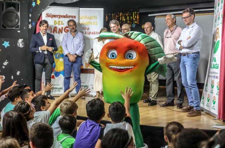 Escolares observan a una mascota con forma de mango junto a varios adultos en un escenario.