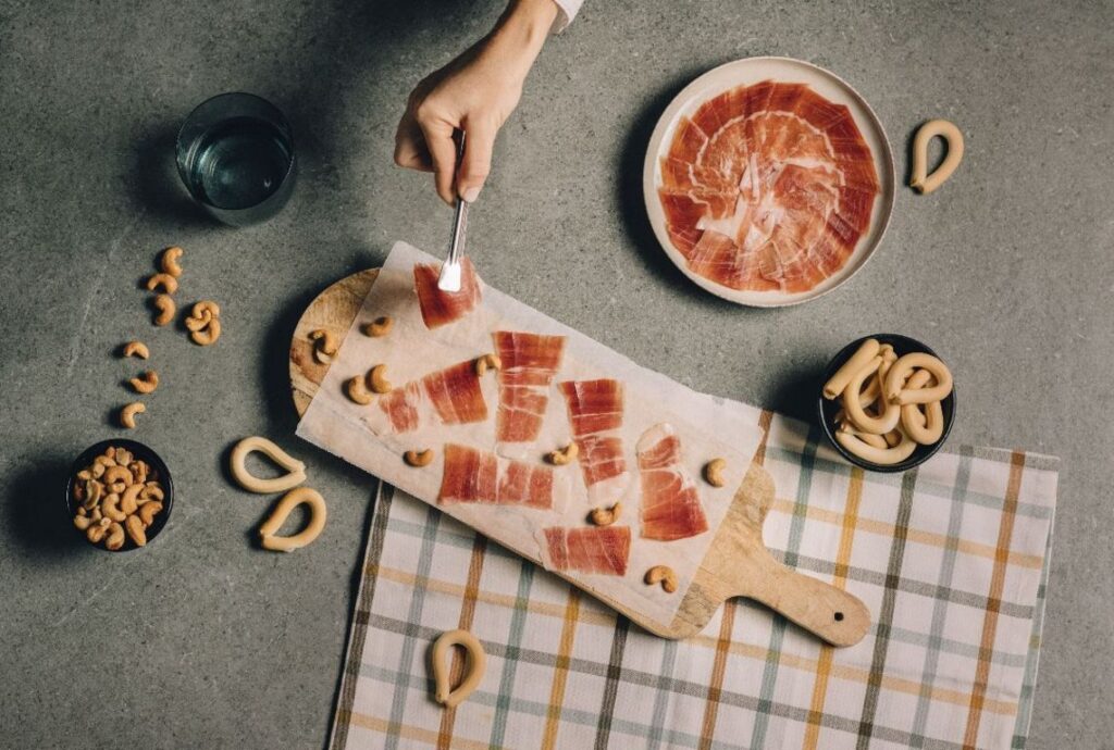 Lonchas de jamón sobre una tabla, con una mano sirviendo una porción y aperitivos alrededor