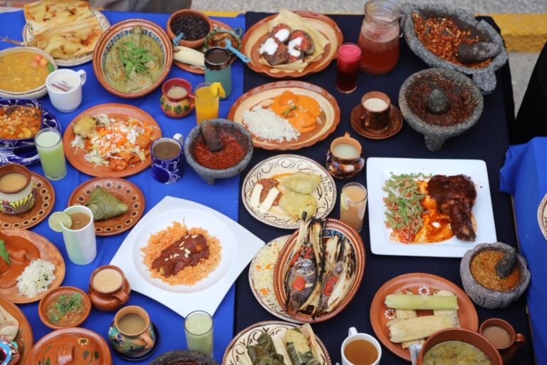 Mesa cubierta de numerosos platos, bebidas y recipientes de cocina sobre manteles azules.