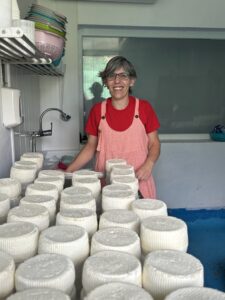 Una mujer sonríe detrás de numerosas piezas redondas de queso blanco apiladas en una sala de trabajo.