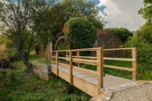 Pasarela de madera junto a un muro de piedra y una estructura circular entre árboles