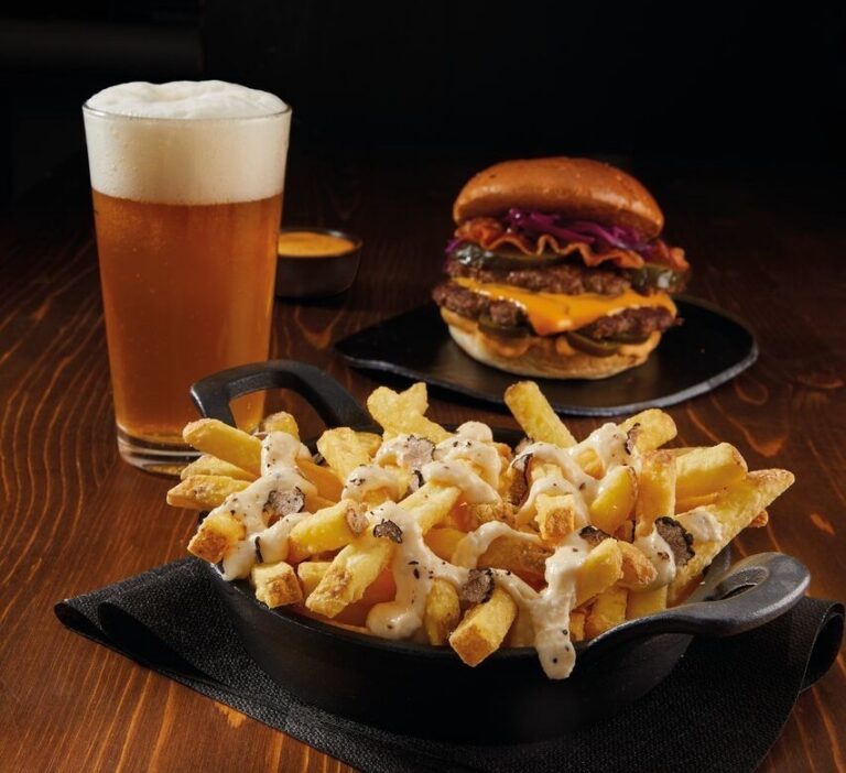Patatas fritas con salsa y trozos oscuros en una sartén, con hamburguesa y cerveza detrás.