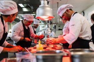 Personas con indumentaria de cocina preparan y emplatan varios platos bajo lámparas de calor