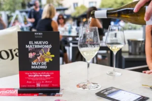 Una mano sirve vino blanco en una copa Rioja junto a un cartel promocional.