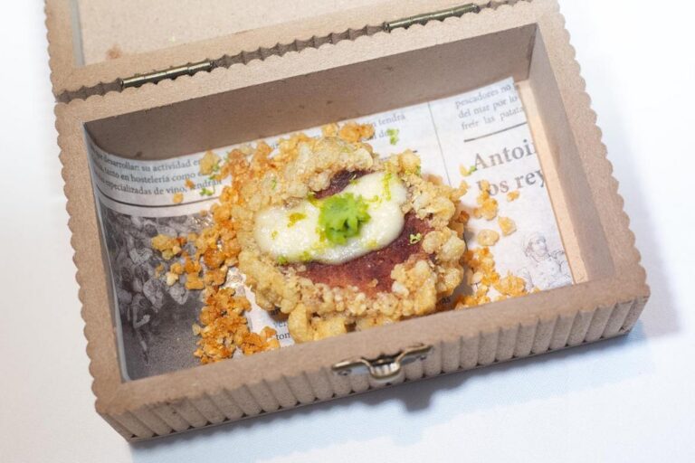 Tapa con base oscura, salsa blanca y crujientes dorados presentada en una caja de cartón.